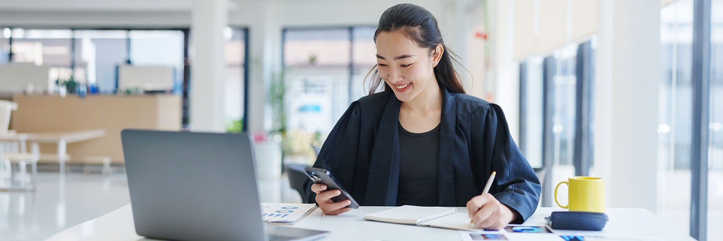 Woman smiling while writing in a notebook and looking at phone; laptop and papers sprawled across desk