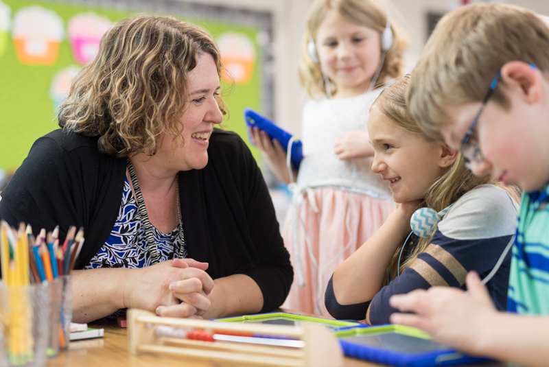 teacher seated at a table, smiling with a group of young students